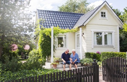 Photo of a yellow cottage with a black roof and an older couple sitting on the porch. A black fence and pink flowers are in the foreground